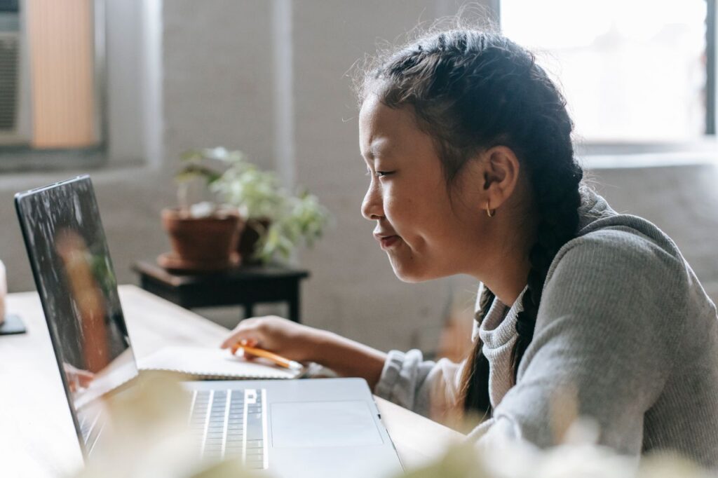 Side view of smart Asian girl sitting at desk and watching laptop in light room